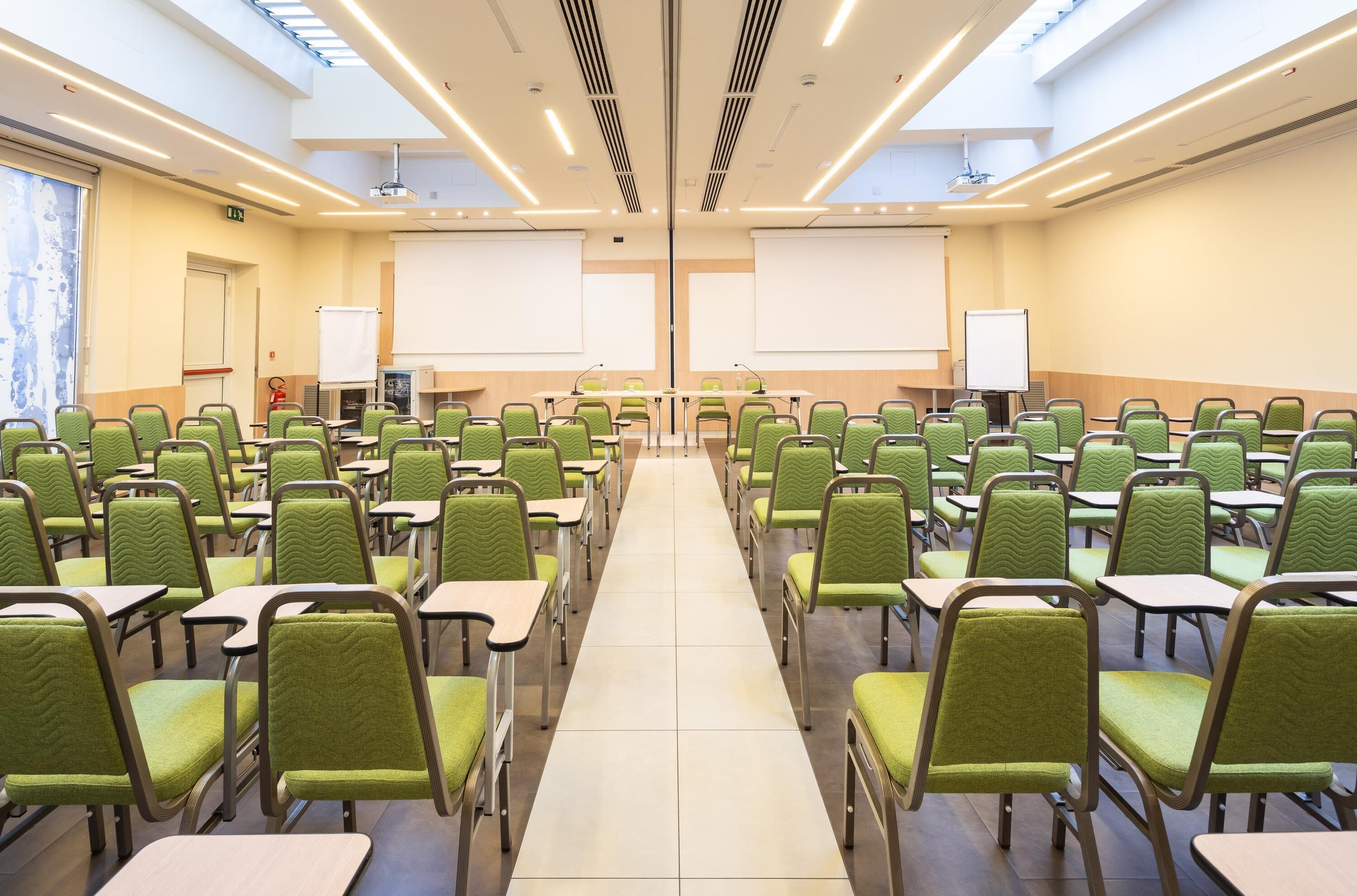 A conference room with rows of green chairs, a long table at the front, and two large white screens.