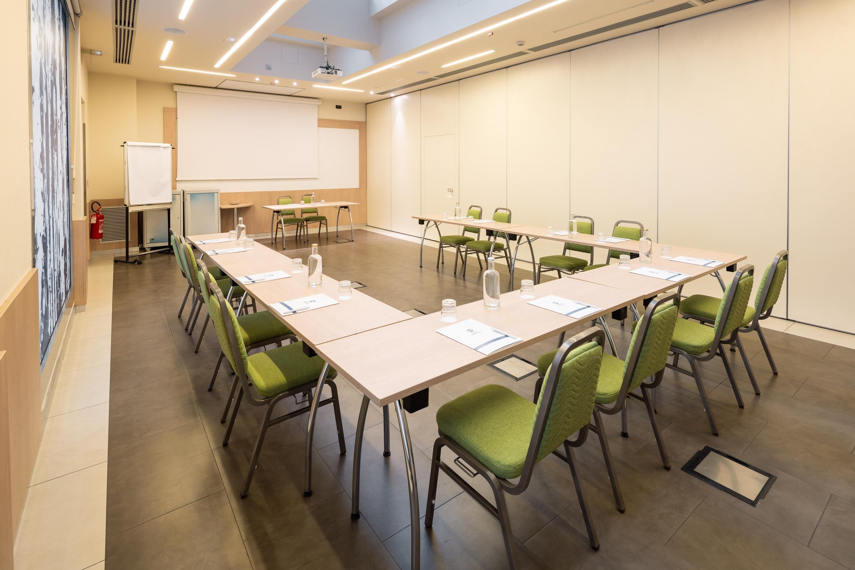 A conference room with a long table, green chairs, a whiteboard, and a projector screen.