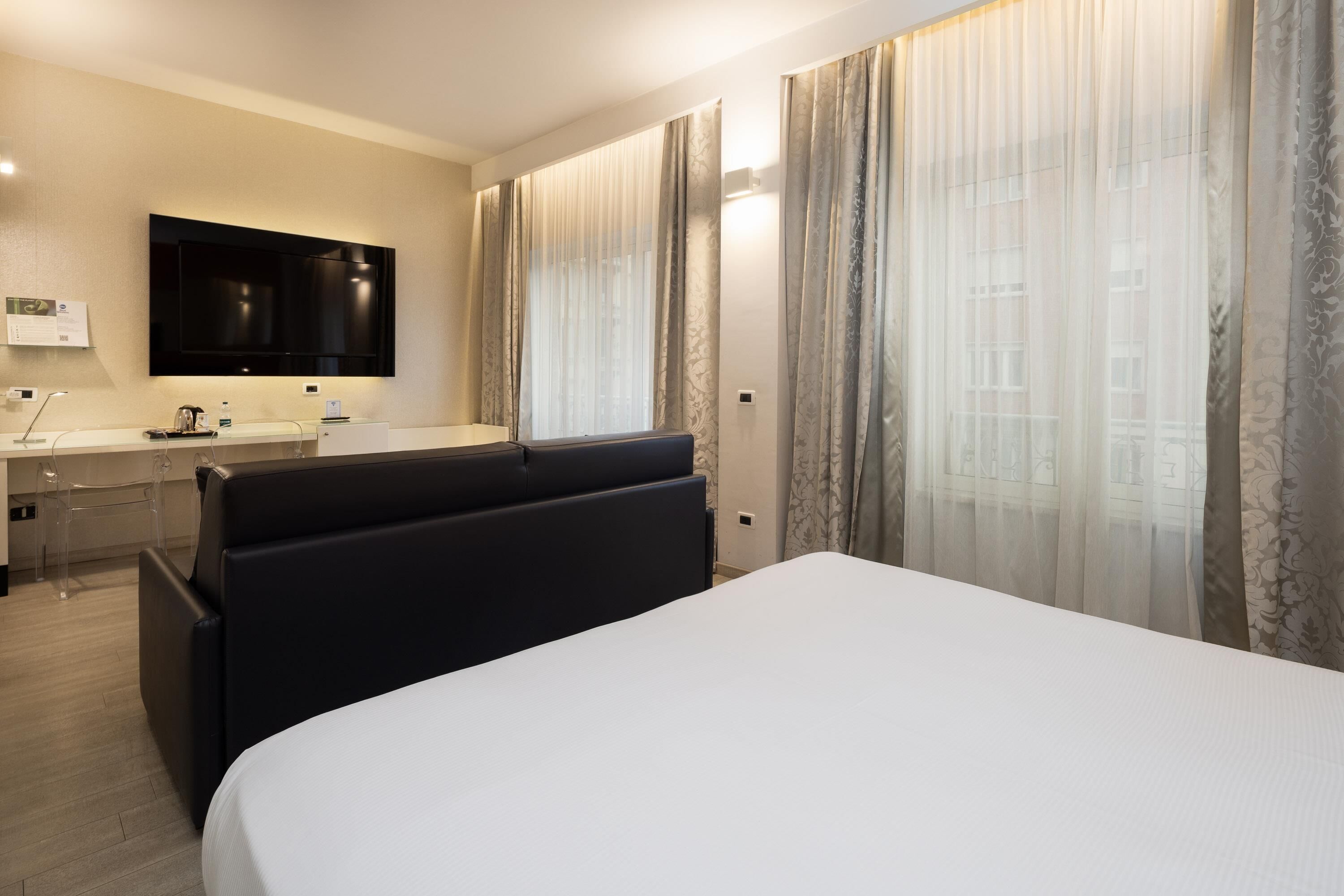 A modern hotel room with a large bed, a flat-screen TV mounted on the wall, a desk with a chair, and a window with sheer curtains.