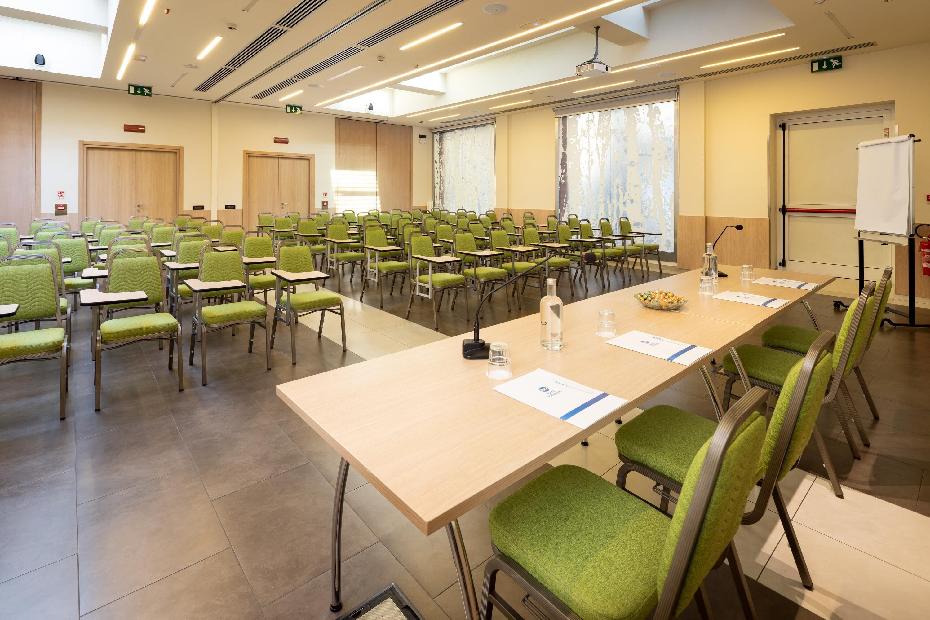 A conference room with green chairs and a long table, equipped with microphones and water bottles.