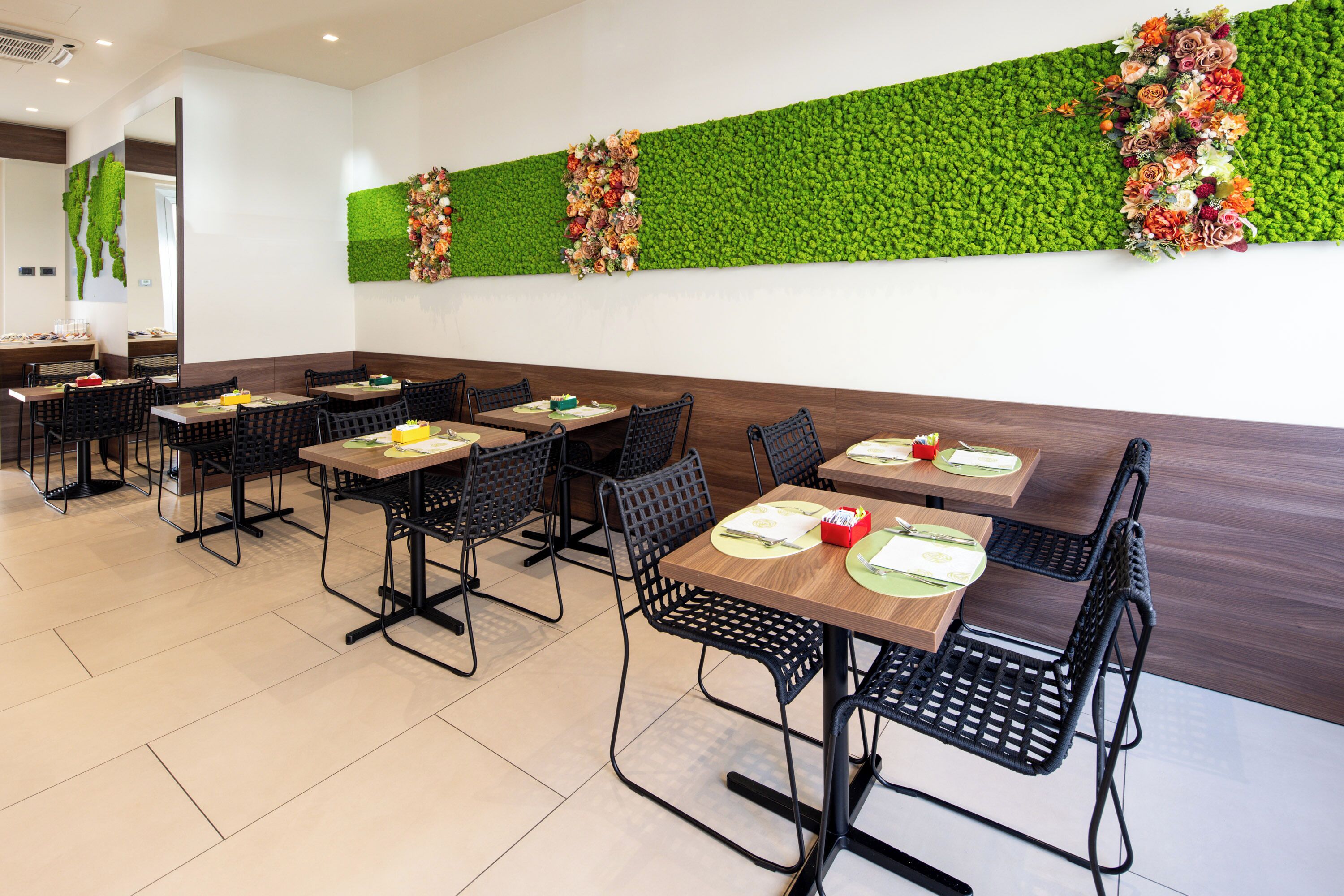 Ein Restaurant mit einer grünen Wand, die mit Blumen geschmückt ist, Holztischen und schwarzen Stühlen.