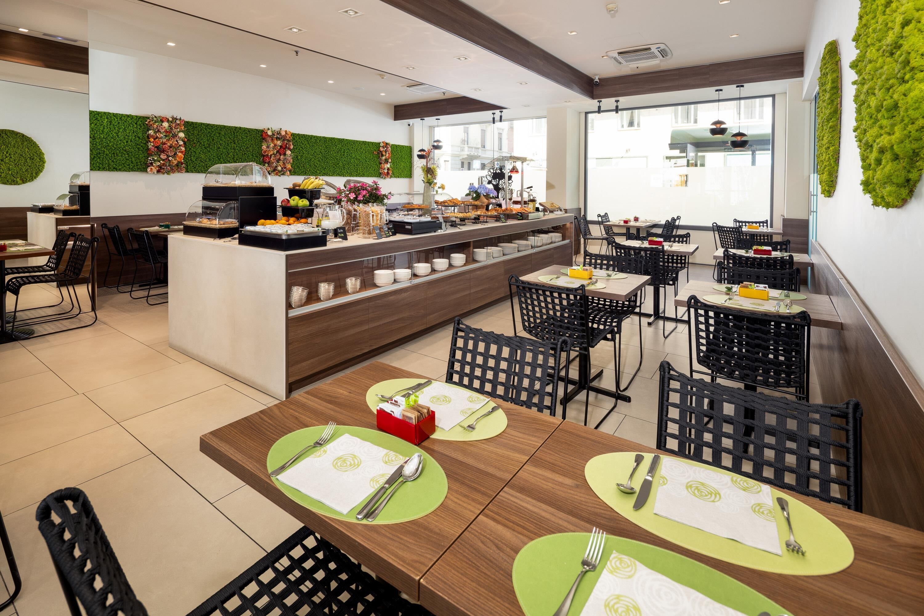 A modern restaurant with a buffet area, multiple dining tables with green placemats, and a green wall with a vertical garden.