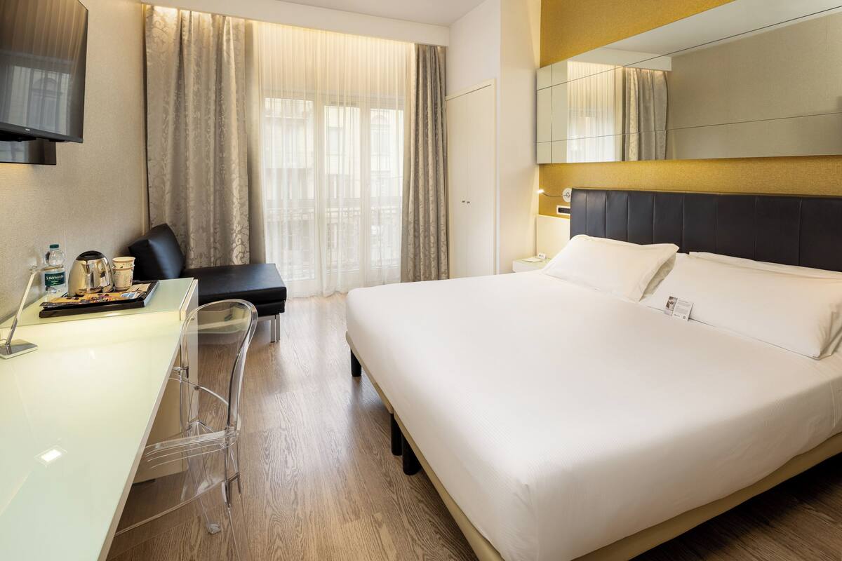 A hotel room with a bed, a desk, a chair, a TV, and a window with curtains.