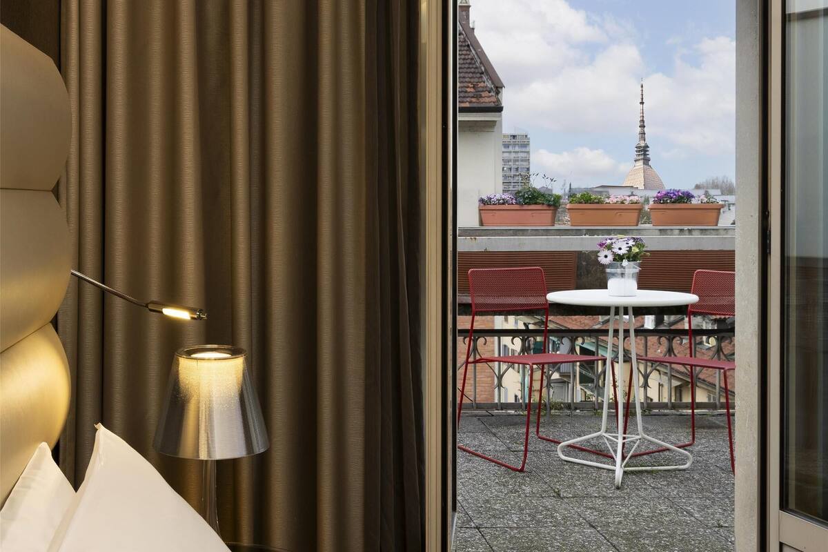 A hotel room with a bed, a lamp, a view of a balcony with a table and chairs, and a cityscape in the background.