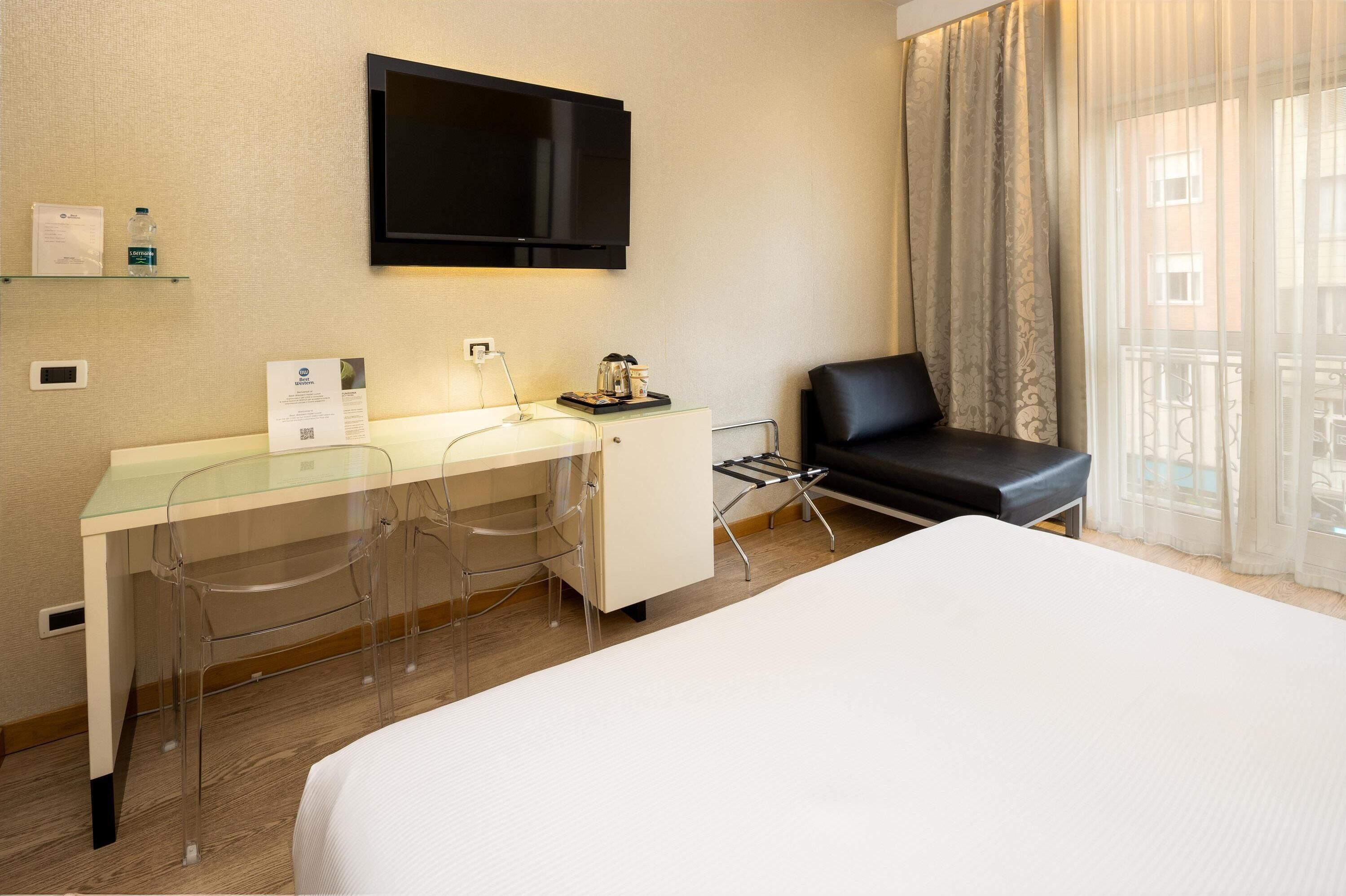 A hotel room with a bed, a desk with a chair, a TV mounted on the wall, and a window with curtains.
