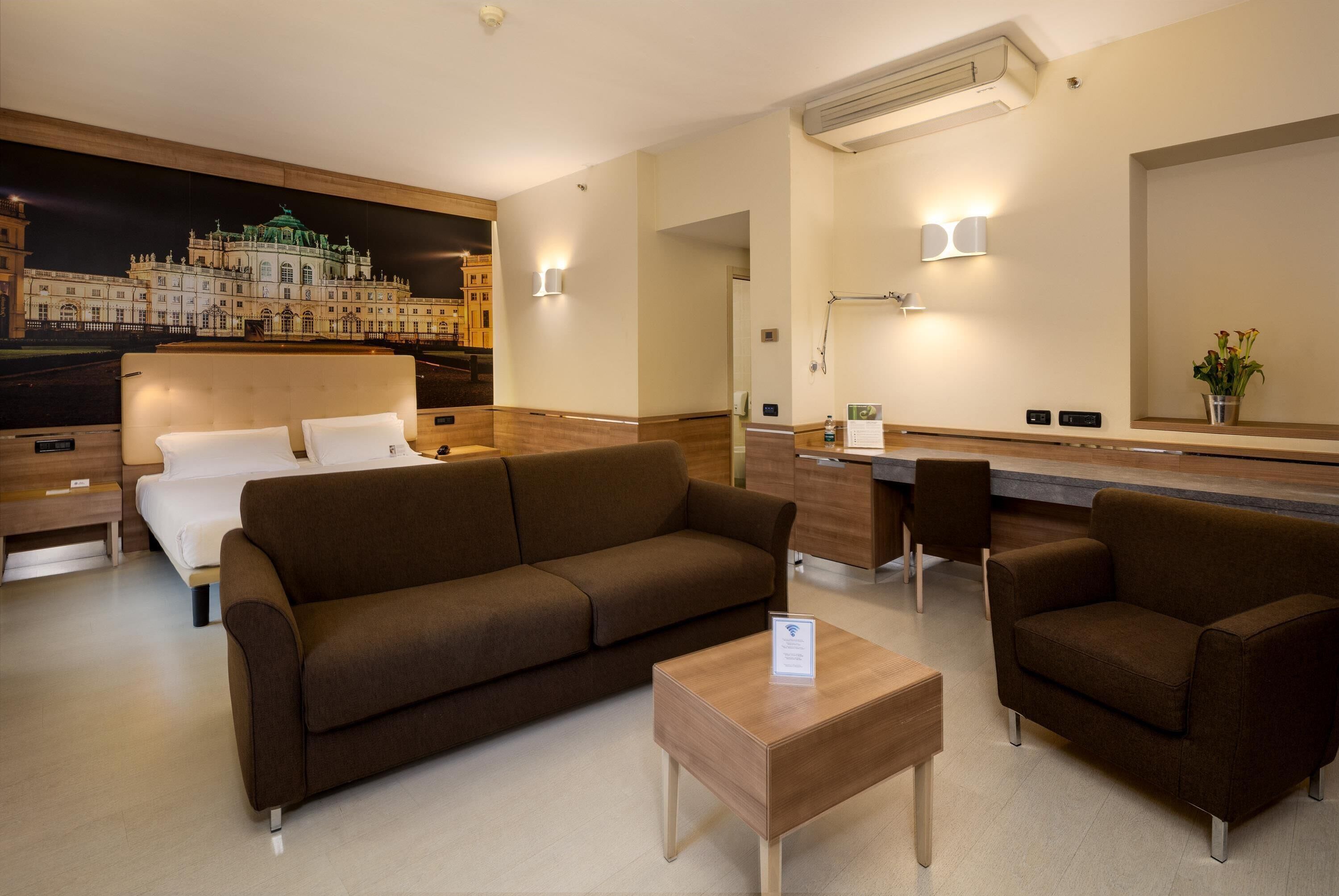 A hotel room with a large bed, a brown sofa, a wooden coffee table, and a desk with a chair. There is a large mirror on the wall and a painting of a building.