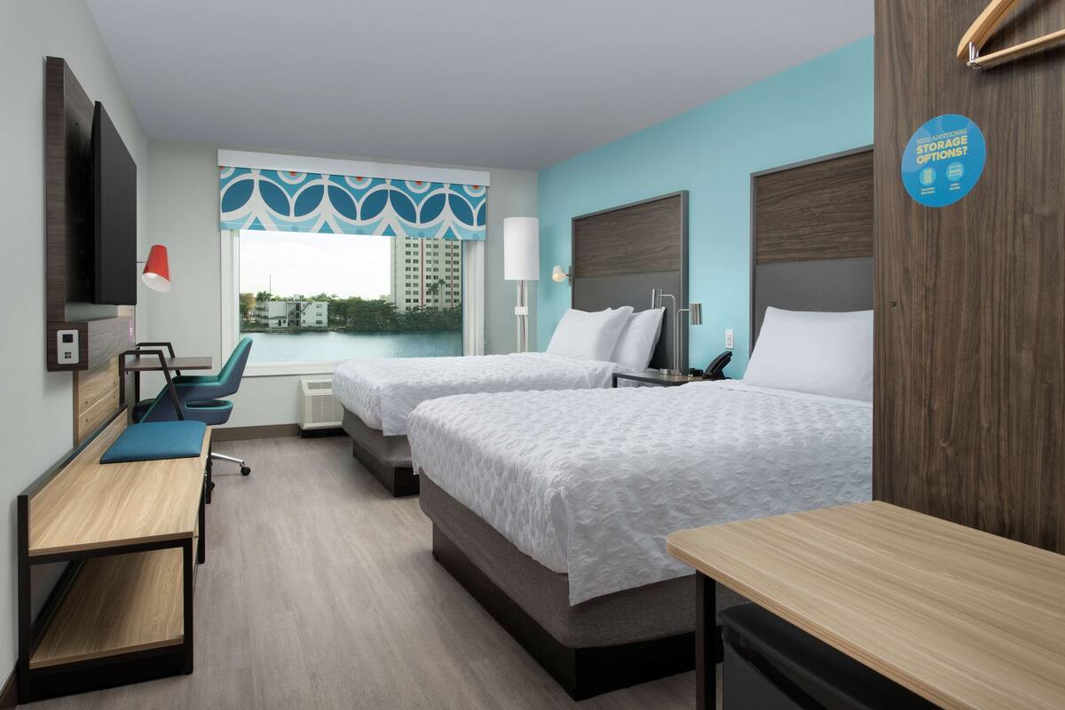 Habitación de hotel con dos camas, un escritorio, una silla, un televisor y una ventana con vista a edificios.