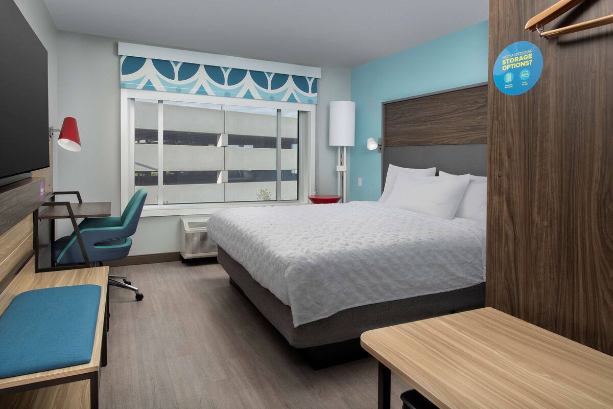 Habitación de hotel con cama, escritorio, silla, televisor y ventana con vista a edificios.