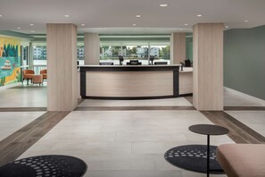 Zona de recepción de hotel con un mostrador de madera, dos sillones, un tapete a cuadrícula en blanco y negro y vistas a edificios a través de amplias ventanas.