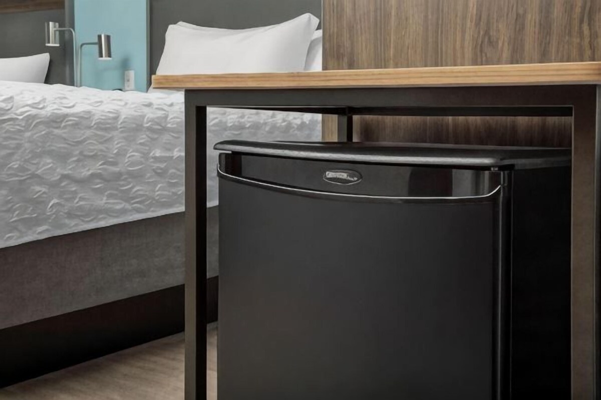 Un refrigerador negro compacto en una habitación de hotel, con una mesa de madera y una cama con cubrecama blanco.