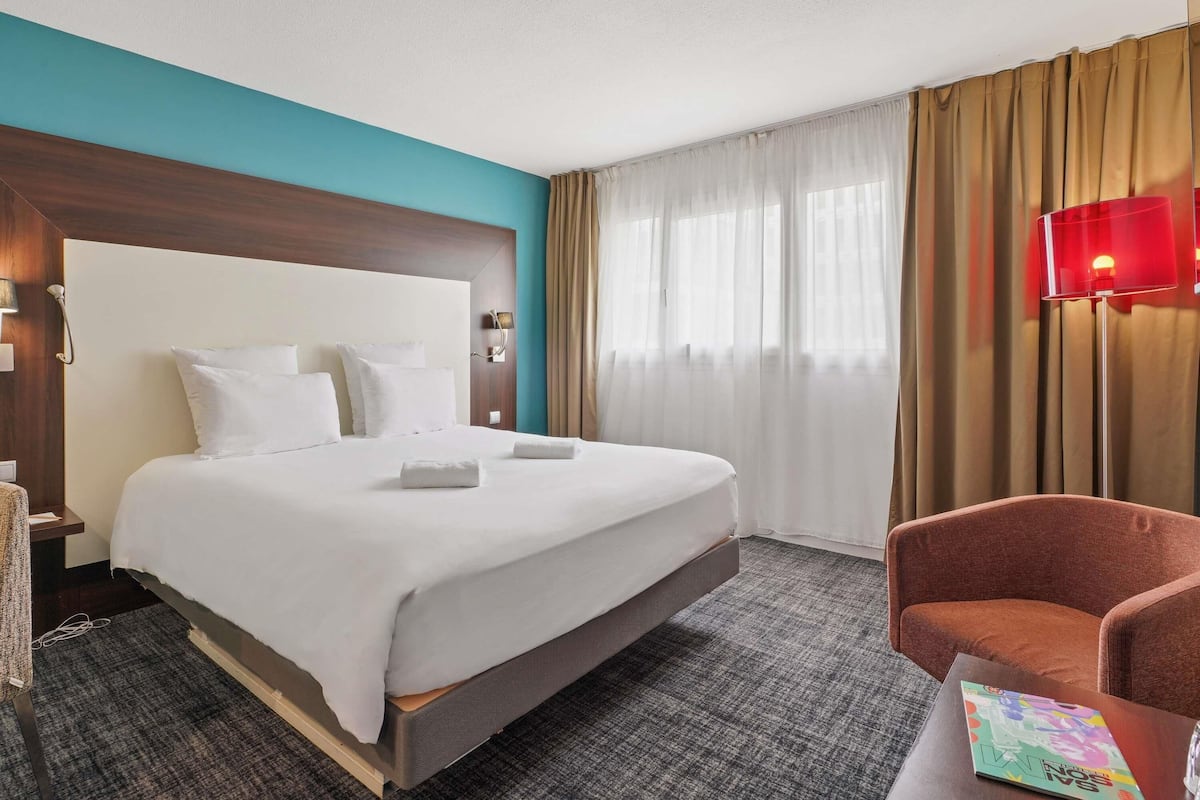 Ein Hotelzimmer mit einem großen Bett, einem Holzkopfende, einer roten Lampe und einem braunen Sessel.