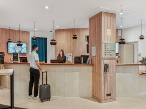 Zona de recepción de un hotel con un mostrador de madera, un televisor, un cartel que enumera los servicios y un hombre con una maleta.