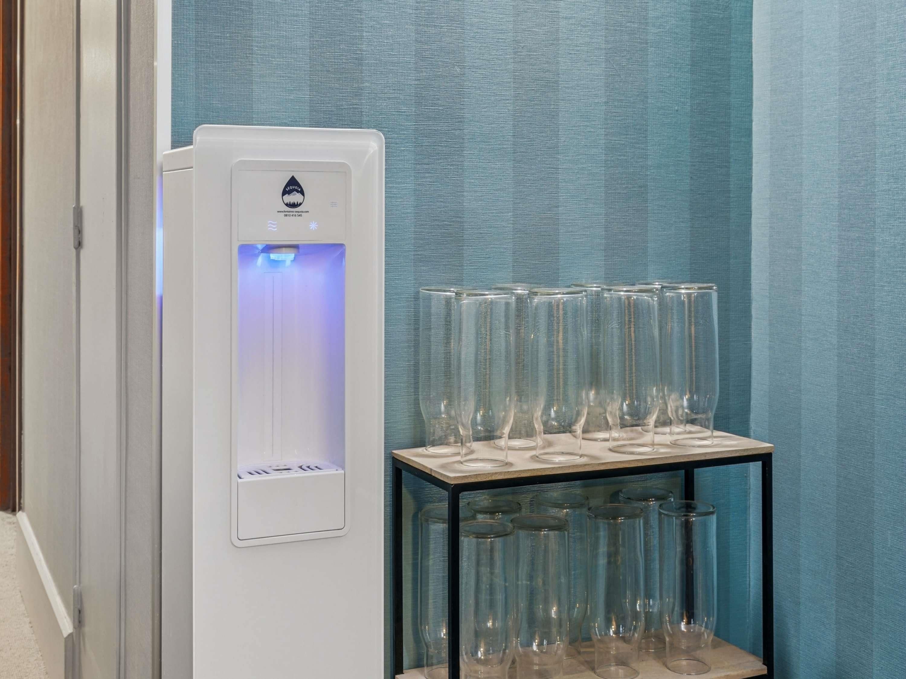 A water dispenser with a logo, a glass display case with multiple glasses, and a blue textured wall.