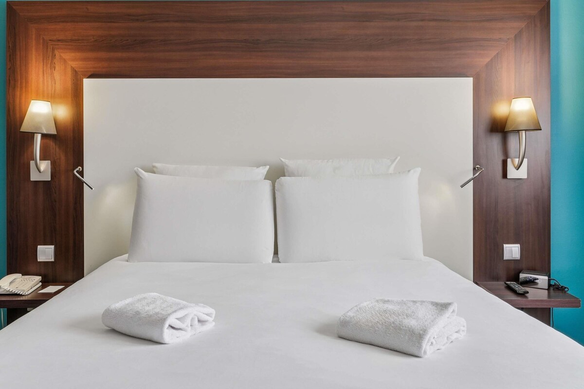 A hotel room with a large bed, two bedside lamps, a telephone, and two towels on the bed.