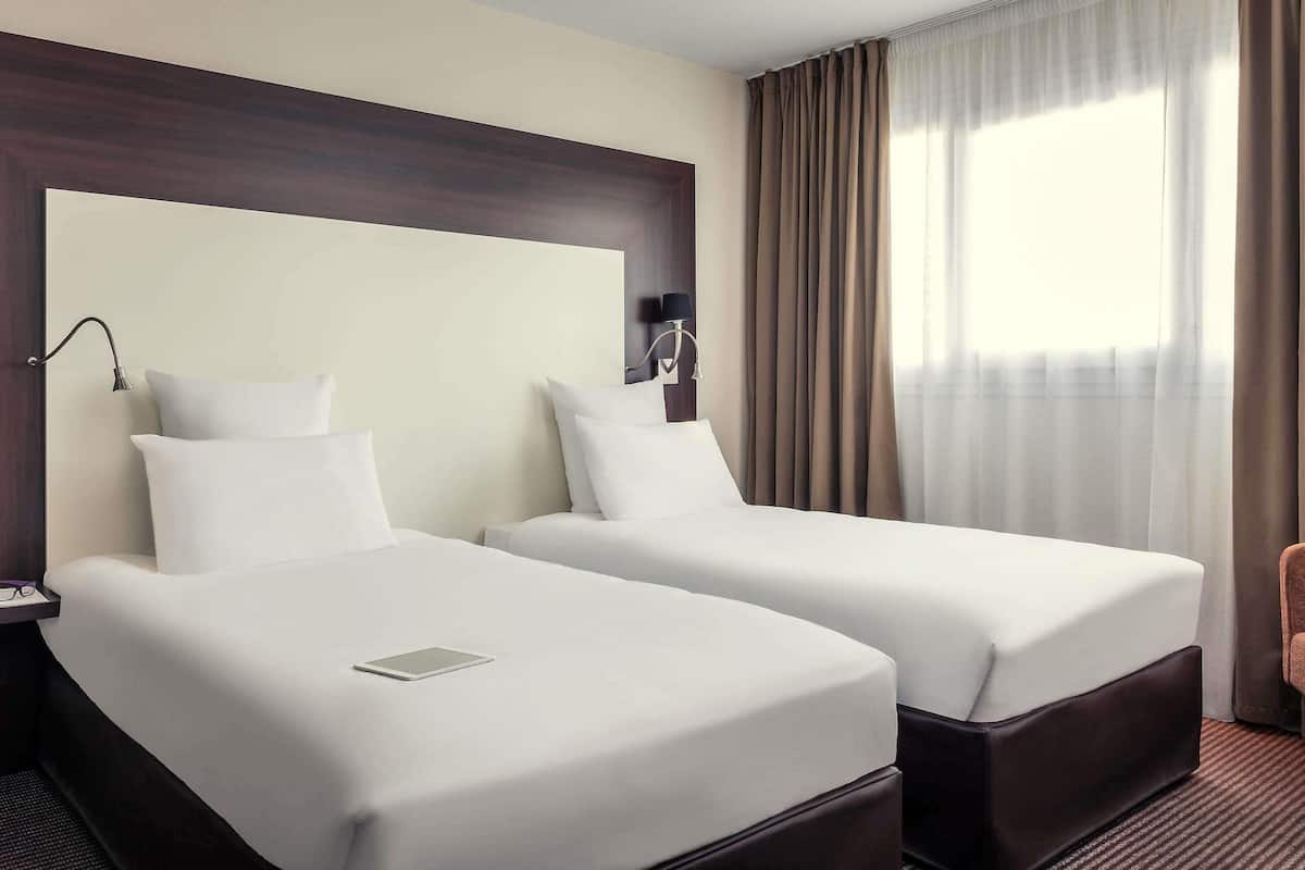 Ein Hotelzimmer mit zwei Betten, einem Nachttisch, einer Lampe und einem Fenster mit Vorhängen.