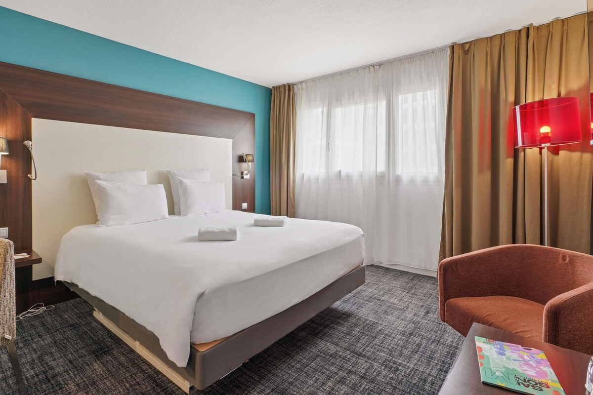 A hotel room with a large bed, a wooden headboard, a red lamp, and a brown armchair.