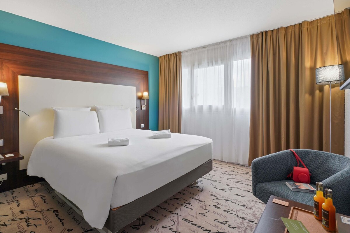 A hotel room with a bed, a desk with a chair, a lamp, and a window with curtains.