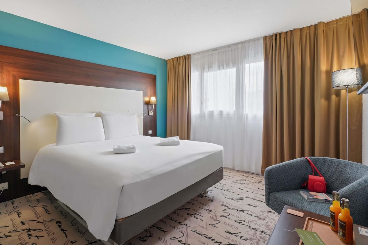 Ein Hotelzimmer mit Bett, Schreibtisch mit Stuhl, Lampe und einem Fenster mit Vorhängen.