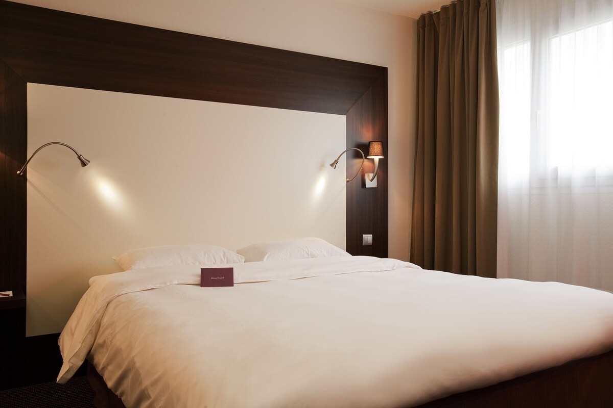 Quarto de hotel com uma cama grande, duas luminárias fixadas na parede e vista para o exterior através de cortinas translúcidas.
