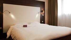 A hotel room with a large bed, two wall-mounted lamps, and a view of the outdoors through sheer curtains.
