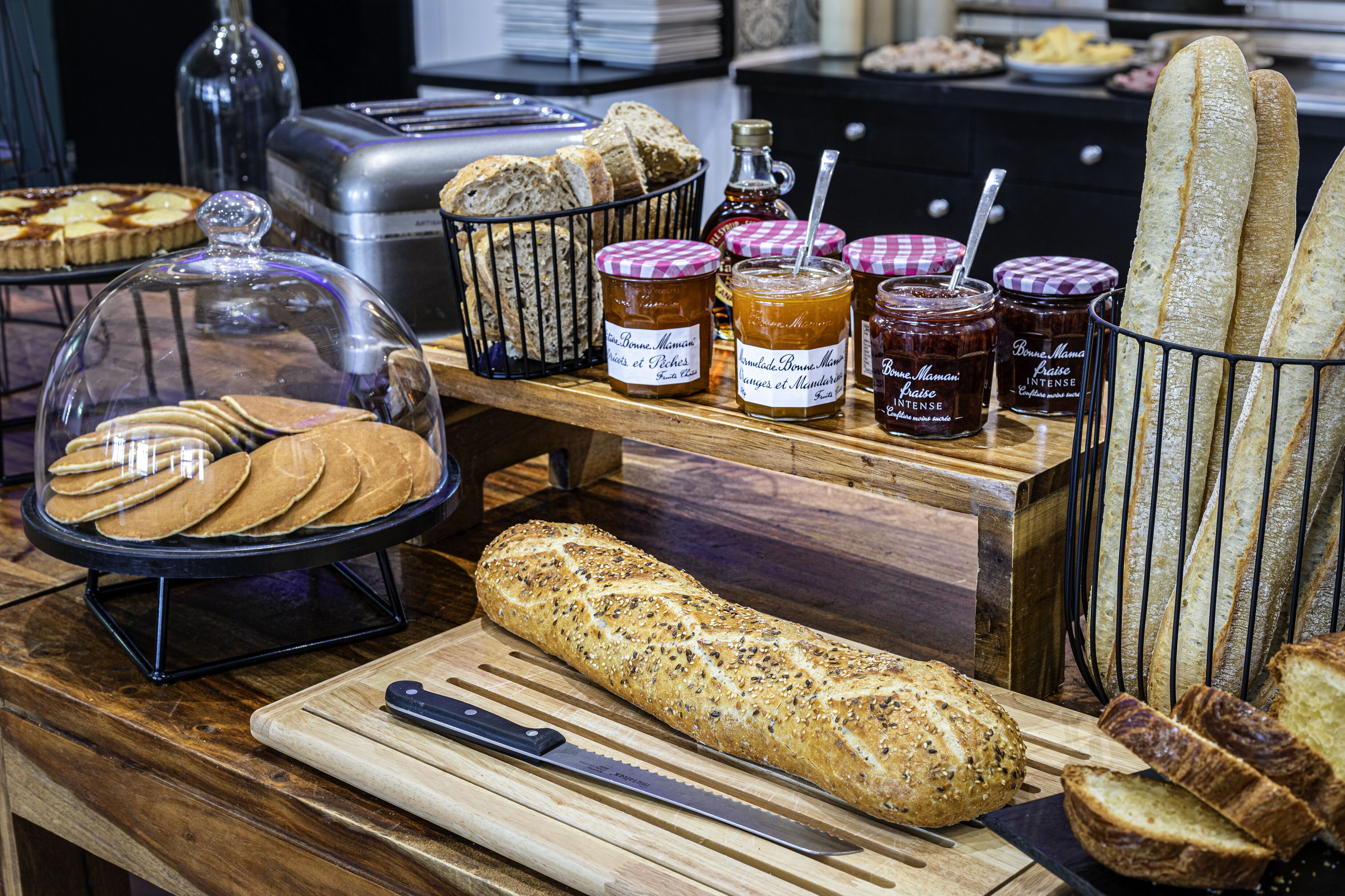 Una vitrina de panadería con diversos panes, una barra de pan con semillas y frascos de mermelada.