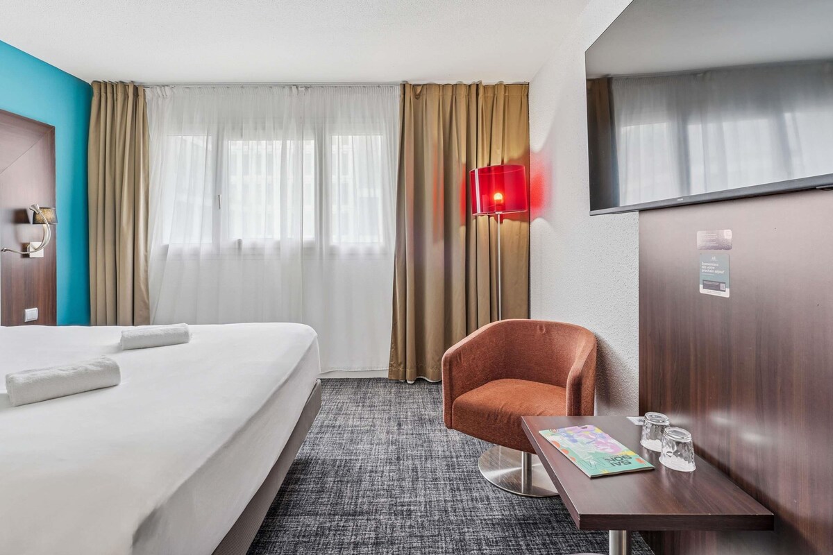 A hotel room with a bed, a chair, a small table with glasses, and a TV.