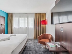 Ein Hotelzimmer mit einem Bett, einem Sessel, einem kleinen Tisch mit Gläsern und einem Fernseher.