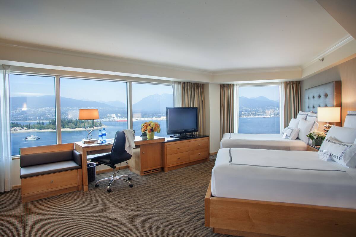 A hotel room with a large window, a desk with a computer, a television, two beds, and a view of a body of water and mountains.