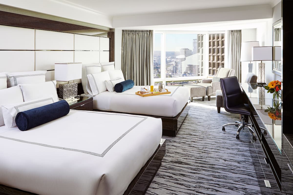 A hotel room with two beds, a desk, a chair, and a large window offering a city view.