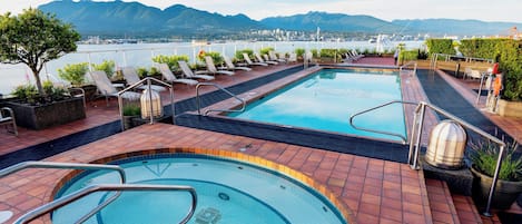 A rooftop pool with two separate sections, surrounded by lounge chairs and potted plants, overlooking a mountainous landscape and a body of water.