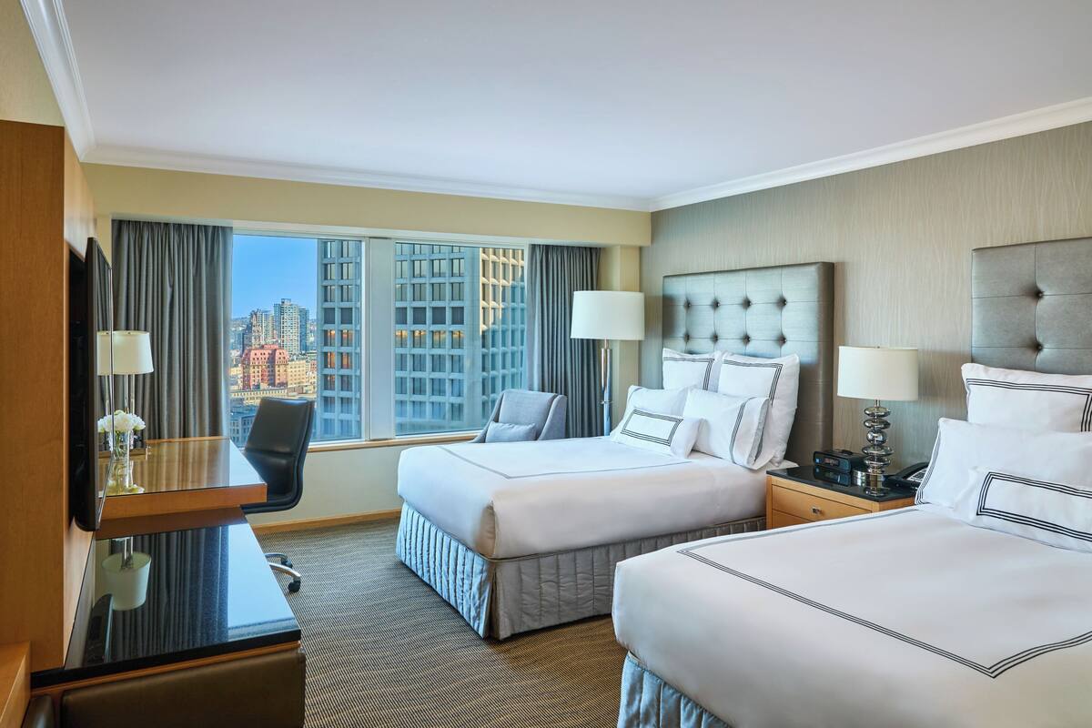 A hotel room with two beds, a desk, and a chair. There is a large window with a city view.