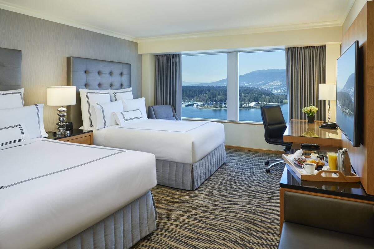 A hotel room with two beds, a desk with a chair, a TV, and a view of a lake and mountains.