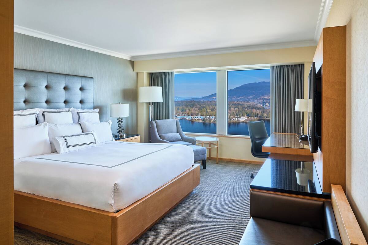 A hotel room with a large bed, a desk, a chair, a TV, and a view of a lake and mountains.