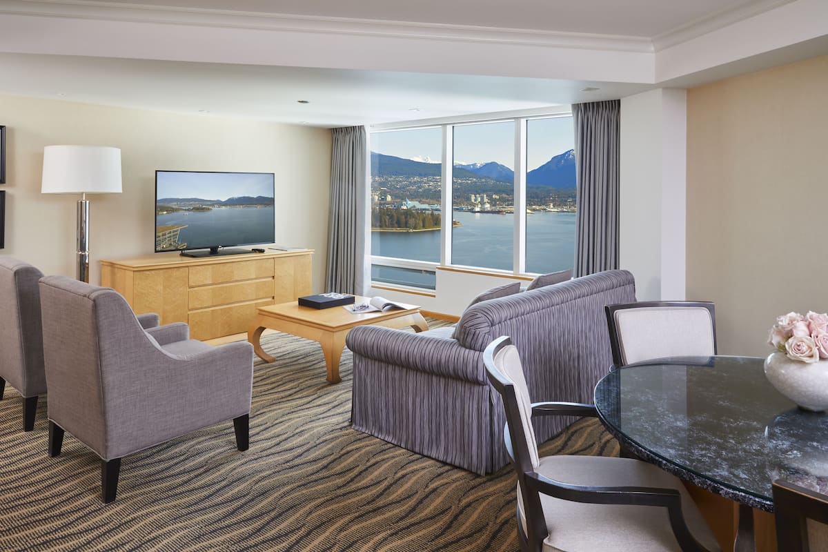 A hotel room with a large window offering a scenic view of mountains and a lake. There is a flat-screen TV on a wooden cabinet, a sofa, a dining table with chairs, and a coffee table.