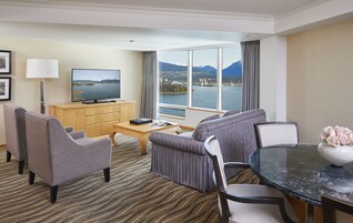 Habitación de hotel con un ventanal grande que ofrece una vista panorámica de montañas y un lago. Hay un televisor de pantalla plana sobre un mueble de madera, un sofá, una mesa de comedor con sillas y una mesa de centro.