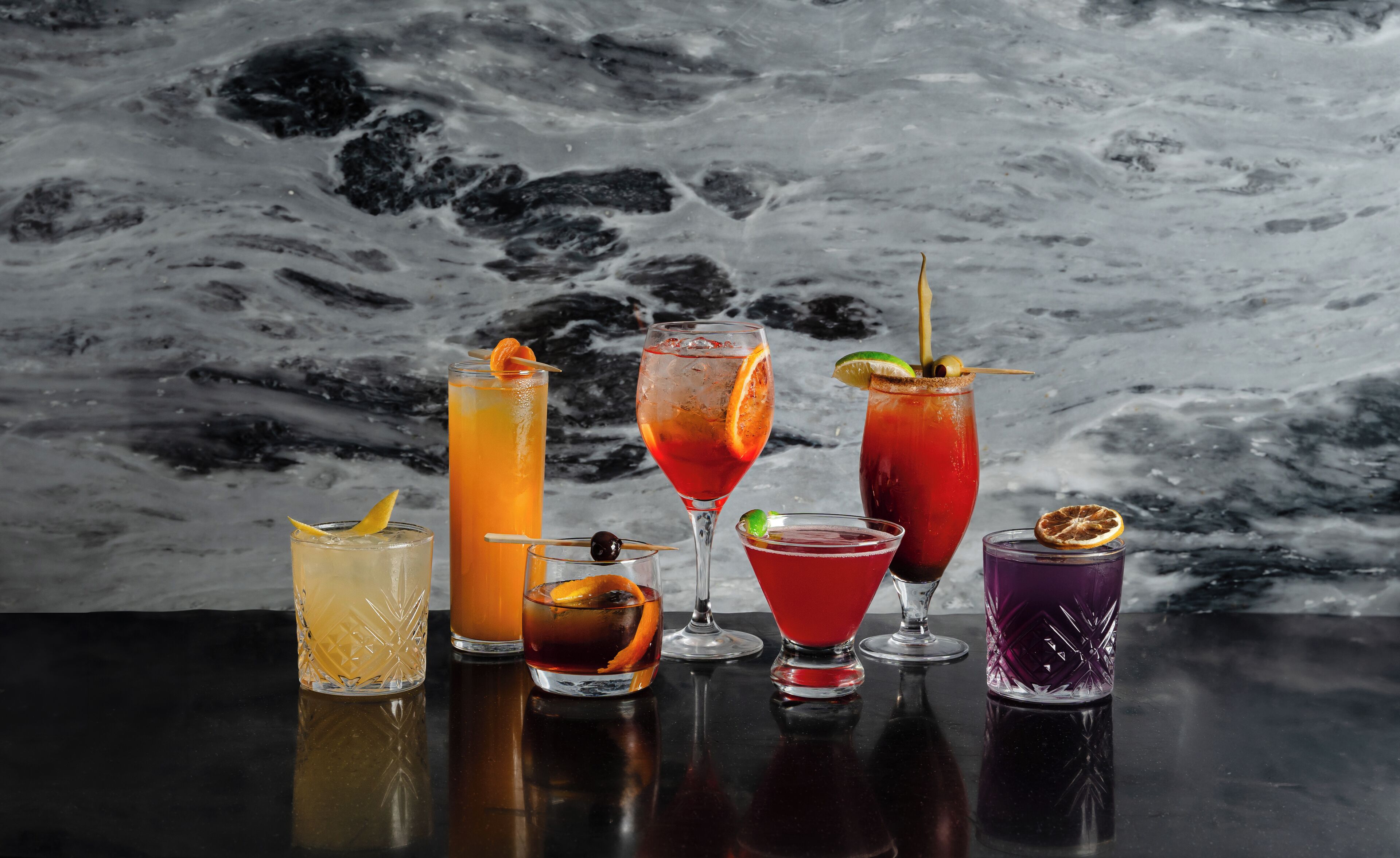 Une sélection de six cocktails présentés sur une surface réfléchissante, avec un fond en marbre.