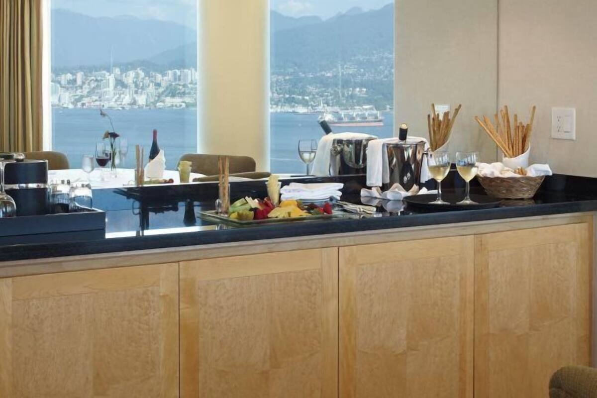 A kitchen counter with a view of a city and water, a fruit bowl, wine glasses, and breadsticks.