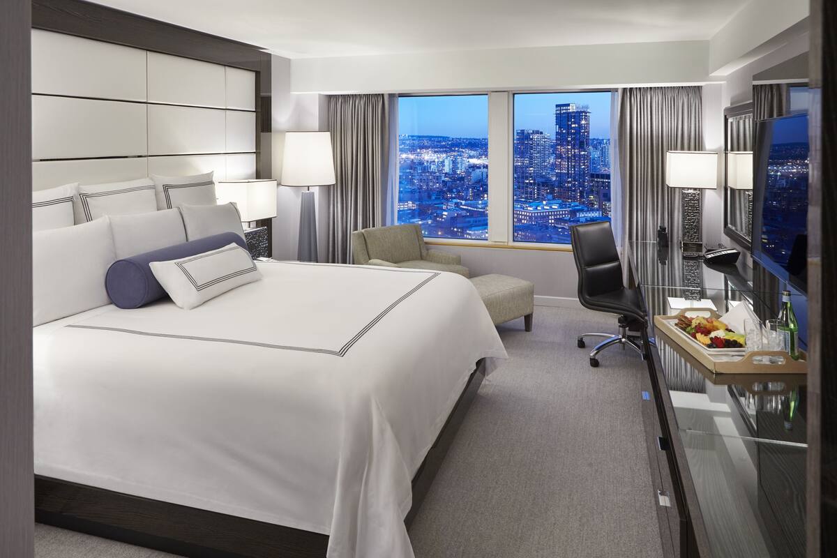 A hotel room with a large bed, a desk with a chair, a TV, and a view of a cityscape.