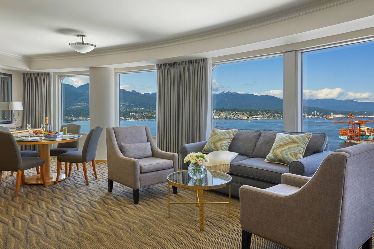 A hotel room with a large window offering a view of a city and mountains, a round dining table with chairs, a sofa, and a coffee table.