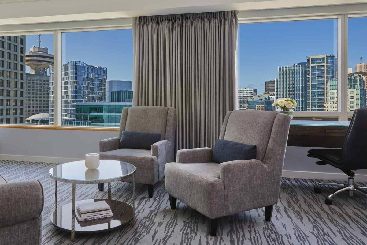 A hotel room with a city view, two armchairs, a sofa, a small table, and a TV.