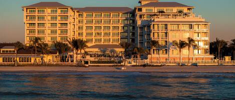 A multi-story hotel with balconies, located near a beach with palm trees.