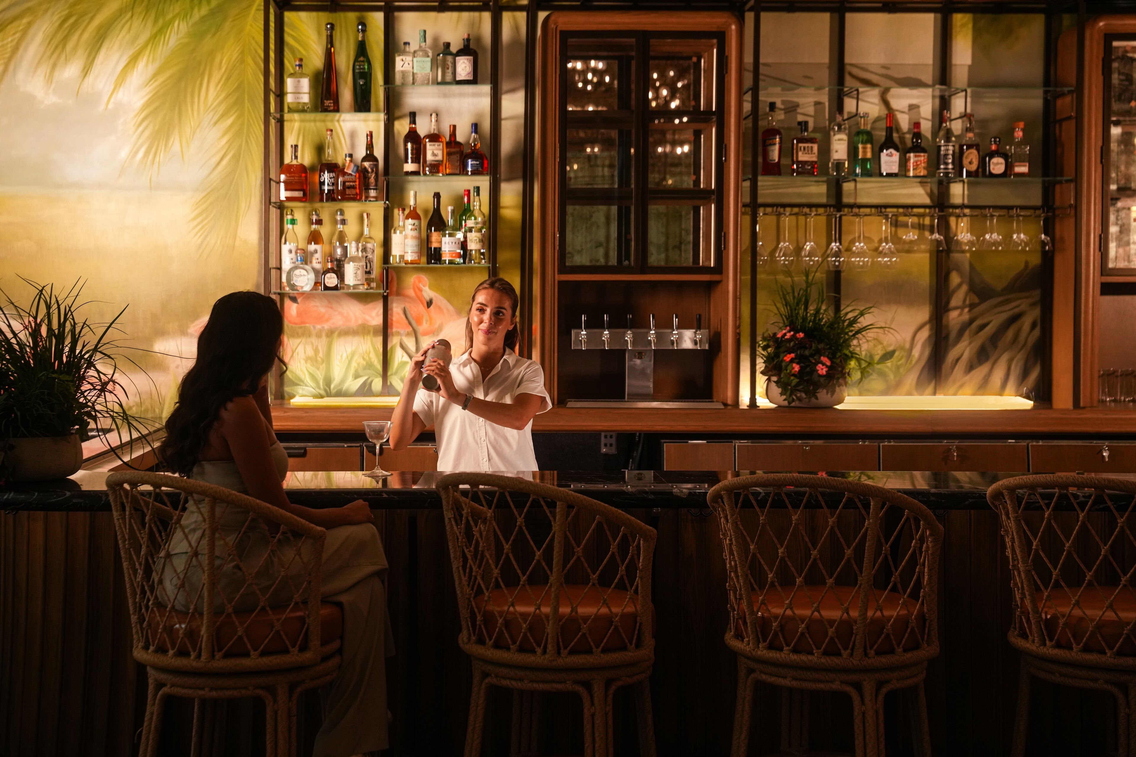 A bar area with wicker chairs, a bartender, and shelves of liquor bottles.