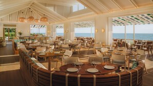 A spacious restaurant with high ceilings and large windows overlooking the sea. Tables are neatly arranged with chairs, and the interior features wooden accents and pendant lighting. - Naples Beach Club, A Four Seasons Resort (Naples)