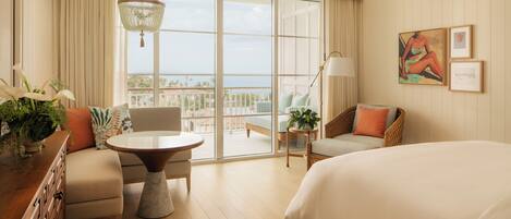 A hotel room with a bed, a sofa, a dining table, and a balcony with a view of the ocean.
