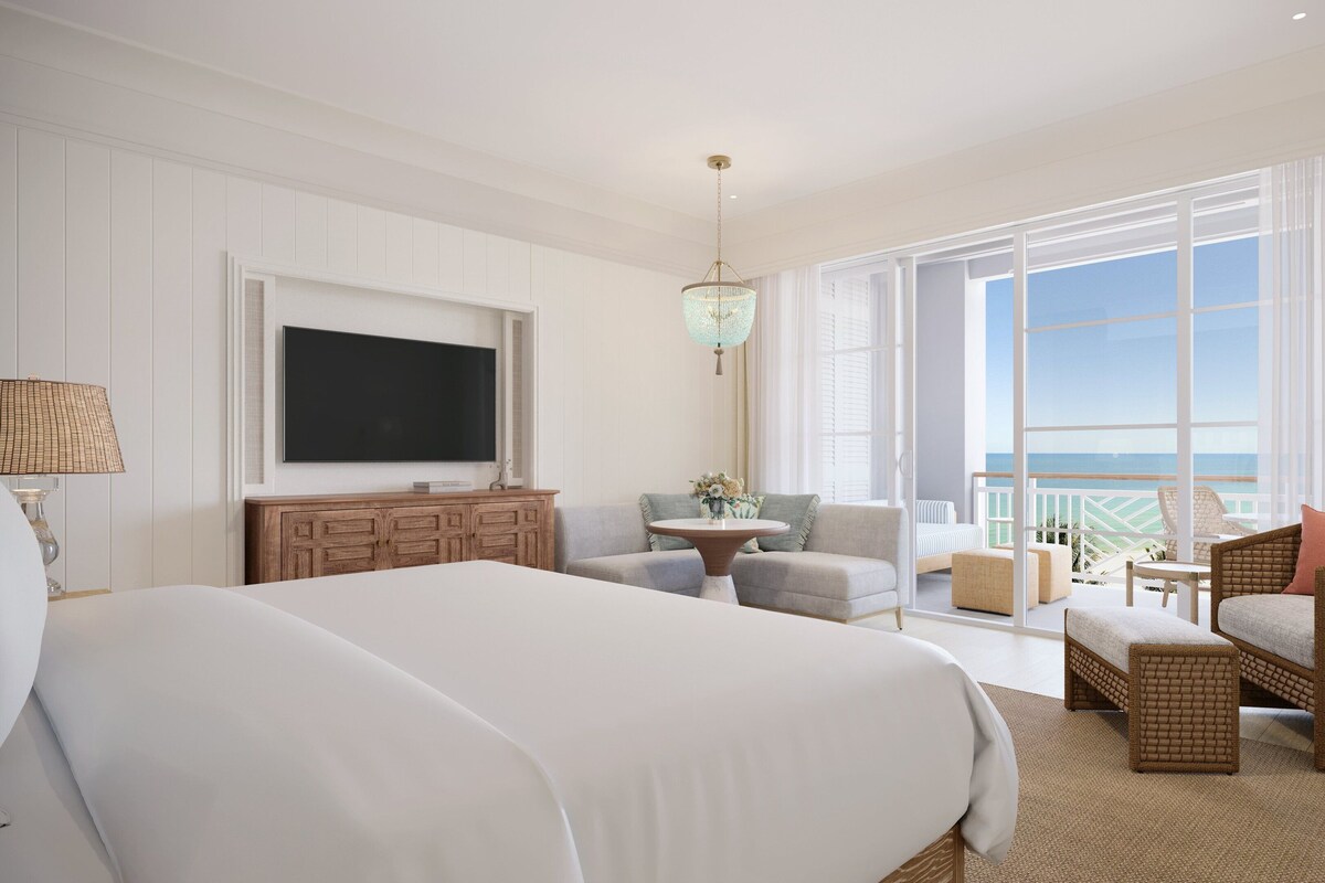 A spacious bedroom with a large bed, a TV, a balcony with a view of the sea, and a wicker chair.