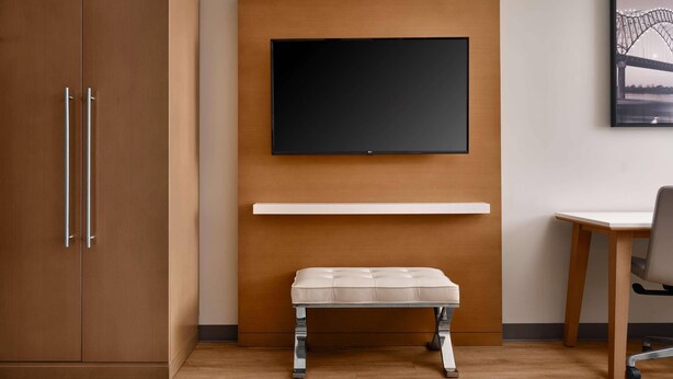 A room with a wooden wardrobe, a flat-screen TV mounted on a wooden panel, a white bench, and a desk with a chair. - Radisson Hotel West Memphis (West Memphis)