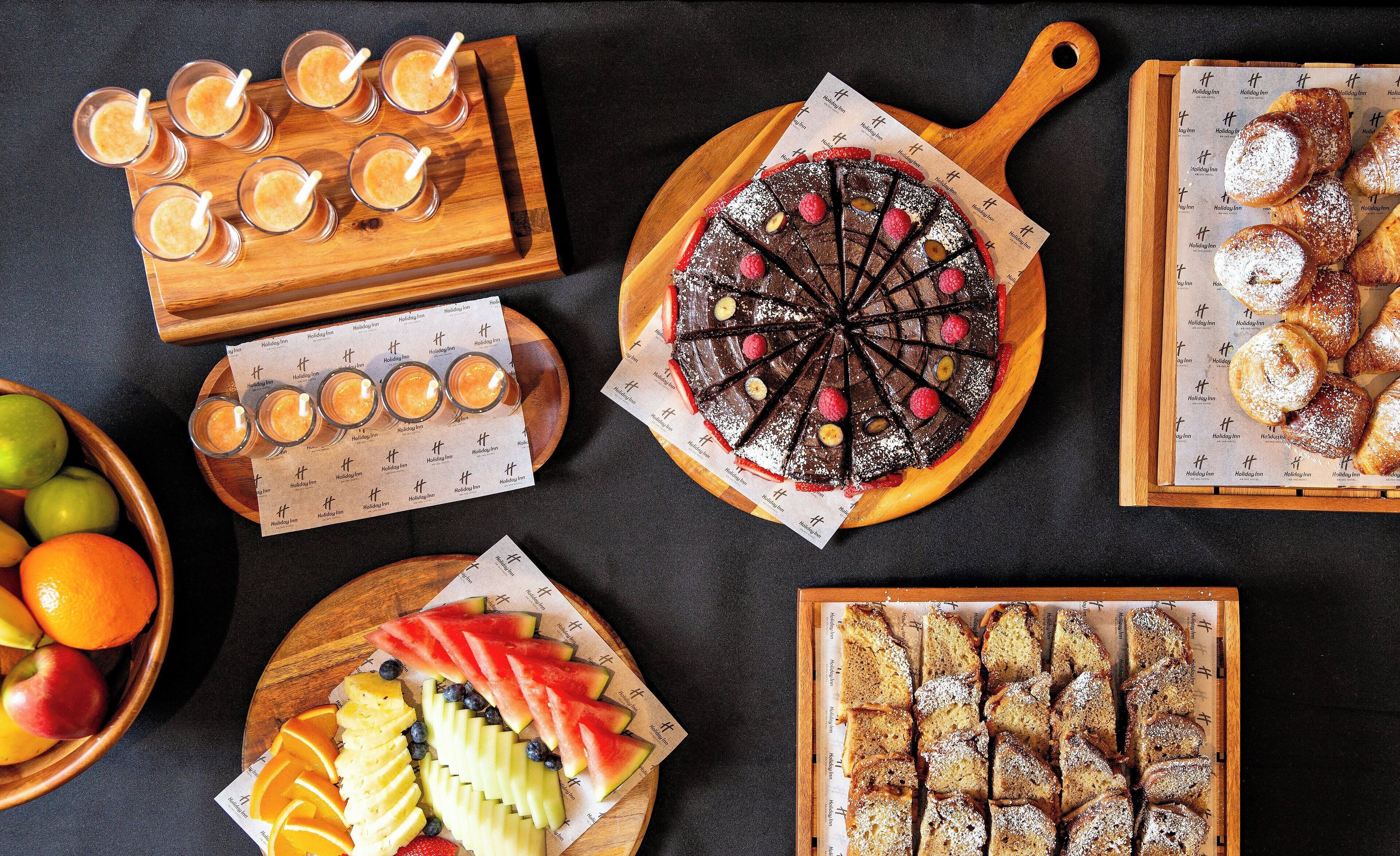 Variedad de alimentos como tarta de chocolate, pasteles, frutas en rodajas y bebidas sobre tablas de madera.