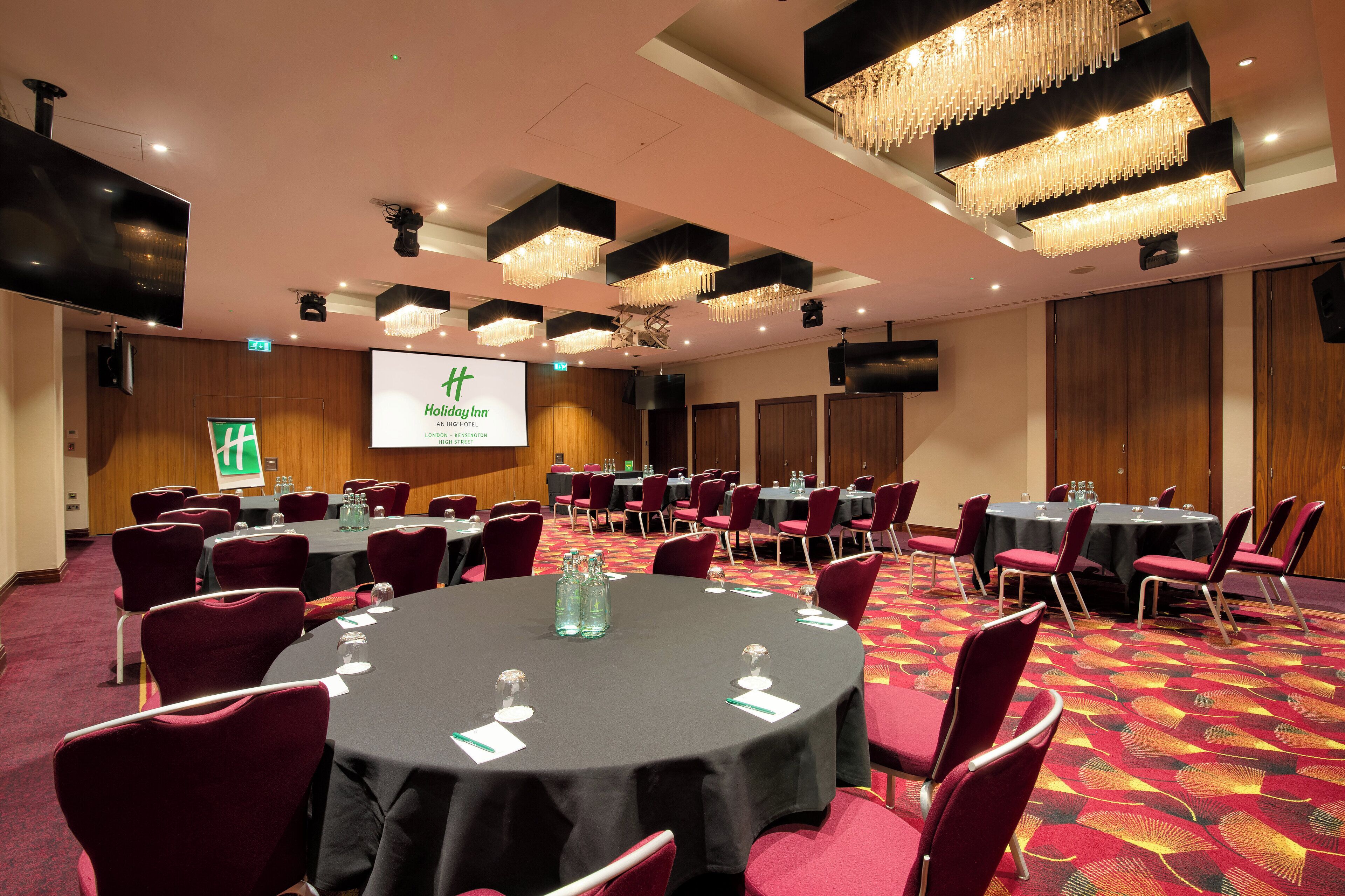 A conference room with round tables, red chairs, and a large screen displaying the Holiday Inn logo.