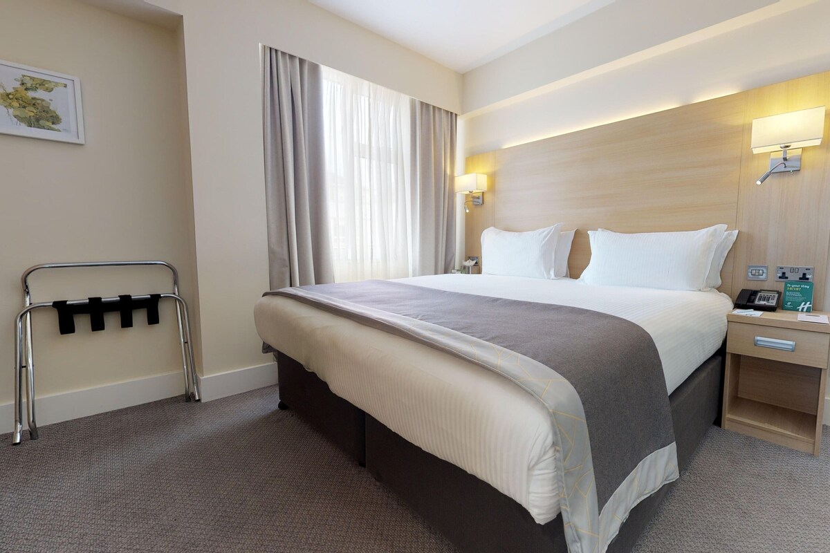 Une chambre d’hôtel avec un grand lit, une table de chevet avec un téléphone et un panneau, un téléviseur posé sur une étagère en bois, et une fenêtre avec des rideaux.