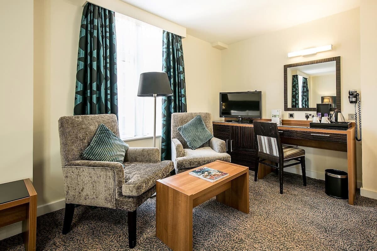 Camera d'albergo con due poltrone, un tavolino in legno, una scrivania con un computer, uno specchio e una TV.